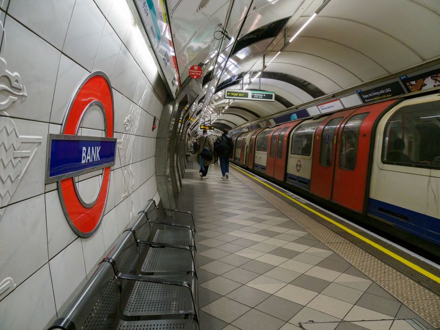 An underground station platform at Green Park with a curved ceiling lined with yellow and white panels. The station walls feature various advertisements and informational posters, including a large ma